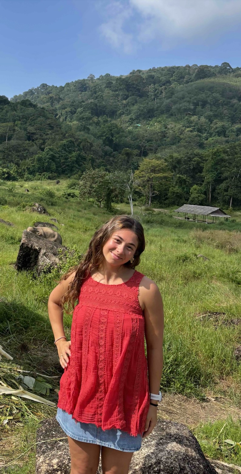 Smiling woman in red top with long hair in a sunny, green mountain landscape.