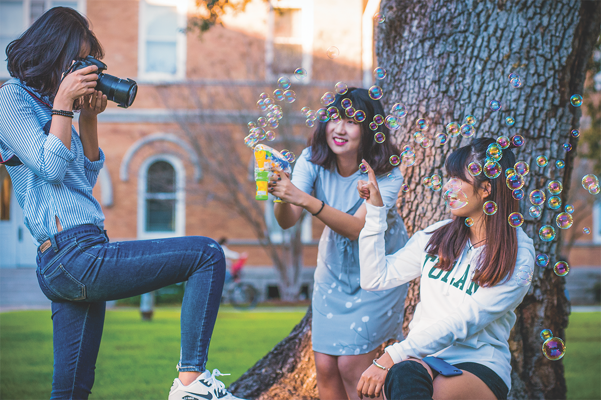 Two students playing with a bubble gun in the quad with a third person taking photos.